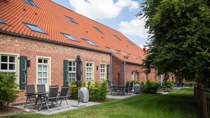 A brick building with a tiled roof and multiple windows. There is a patio area with outdoor seating and a covered grill. - Traumhafte Fewo mit Terrasse im Historischen Hof - Gulfhof Boomgaarden (Krummhörn)