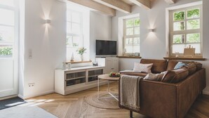 A modern living room with a wooden ceiling, white walls, a brown leather sofa, a TV, and a small table with a bowl of fruit. - Traumhafte Fewo mit Terrasse im Historischen Hof - Gulfhof Boomgaarden (Krummhörn)