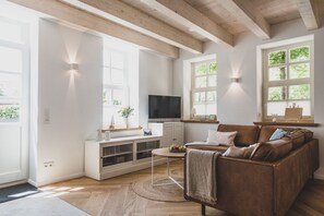 Een moderne woonkamer met een houten plafond, witte muren, een bruine leren bank, een televisie en een kleine tafel met een schaal fruit.