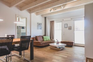 A spacious living area with a wooden floor, a dining table with chairs, a sofa with cushions, a coffee table, and a large window with curtains. - Moderne und Gemütliche Fewo "gerjet" - Gulfhof Boomgaarden (Krummhörn)