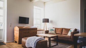 Ein Wohnzimmer mit braunem Ledersofa, einer hölzernen Kommode, einem Couchtisch, einer Stehlampe und einem an der Wand befestigten Fernseher.