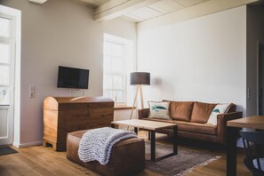 A living room with a brown leather sofa, a wooden chest of drawers, a coffee table, a floor lamp, and a television mounted on the wall. - Modern und Gemütlich "mettinus" - Gulfhof Boomgaarden (Krummhörn)