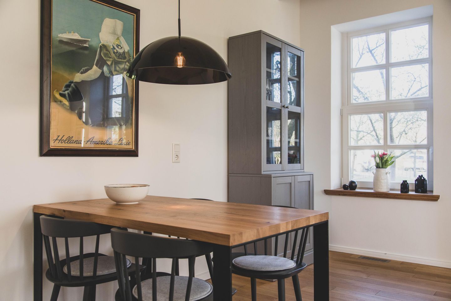 A dining area with a wooden table, black chairs, a black pendant light, a cabinet, and a window with a view of trees.