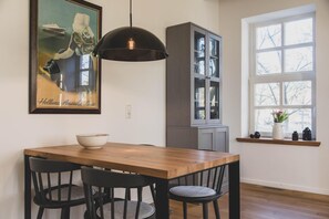 Uma sala de jantar com mesa de madeira, cadeiras pretas, luminária pendente preta, um armário e uma janela com vista para árvores.