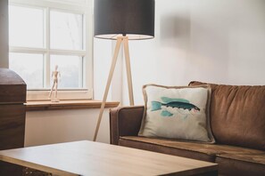 Ein Wohnzimmer mit braunem Ledersofa, einem Holztisch, einer Stehlampe und einem Fenster mit Blick auf Bäume.