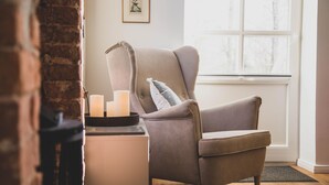 Ein gemütliches Zimmer mit einem grauen Sessel, einem kleinen Tisch mit Kerzen und einem Fenster mit Blick auf Bäume.