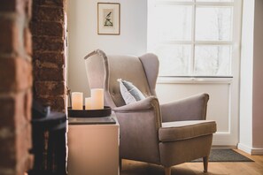 A cozy room with a grey armchair, a small table with candles, and a window with a view of trees. - Modern und Gemütlich "mettinus" - Gulfhof Boomgaarden (Krummhörn)