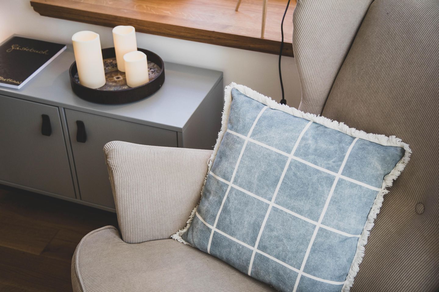 A chair with a blue and white checkered pillow, a grey upholstered armchair, and a small side table with three white candles and a book titled "Jisabook".