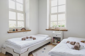 A bright, minimalist room with two single beds, a small desk, and a window with a view of greenery.