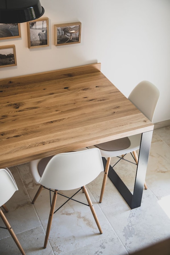 A wooden dining table with four white chairs, a black hanging lamp, and three framed pictures on the wall.