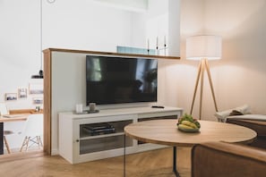 A modern living room with a flat-screen TV, a round wooden table with a fruit bowl, a standing lamp, and a white entertainment unit.