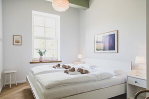 A hotel room with two beds, a window with a view, a bedside table with a lamp, and a framed picture on the wall.