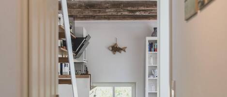 A hallway with wooden flooring, a staircase, a bookshelf, and a window with a view of greenery.