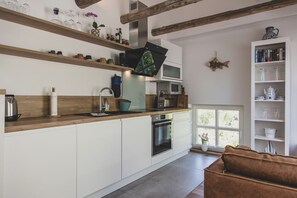 Une cuisine moderne avec des meubles blancs, des plans de travail en bois et un four encastré. Une fenêtre apporte de la lumière naturelle et une bibliothèque est agrémentée d’objets décoratifs.