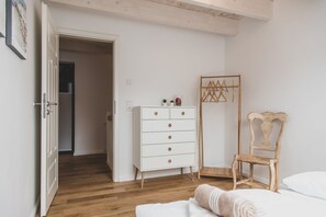 Un dormitorio con suelo de madera, cómoda blanca, silla de madera y cama con ropa de cama blanca.