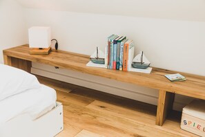 A neatly organized bedroom with a wooden bed frame, a bedside table with a white lamp, a small bookshelf, and a white storage box labeled "SPIEL".