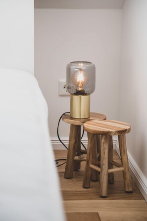 Ein kleiner Holzschemel mit einer Tischlampe darauf, eine weiße Wand und eine Steckdose im Hintergrund.