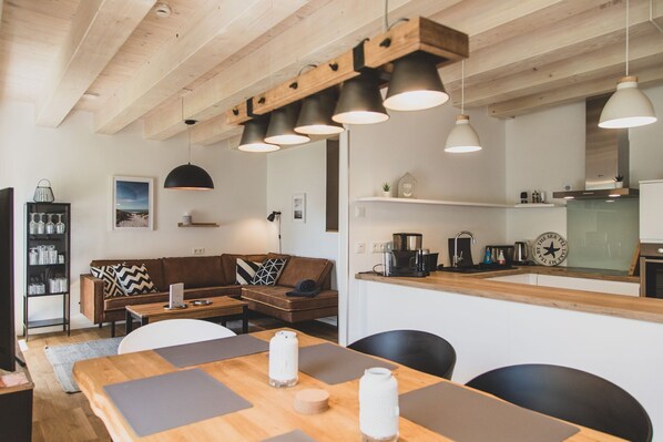 A modern living room with a wooden ceiling, a brown sofa, a dining table with mats, and pendant lighting.
