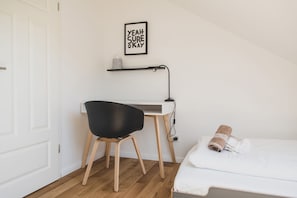 A small, neatly organized bedroom with a single bed, a desk with a chair, and a wall-mounted shelf.