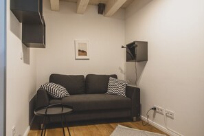 A small, modern living room with a dark grey sofa, two patterned pillows, a small round table, and a single framed picture on the wall. - Exklusive Maisonette Ferienwohnung "aafke" im Gulfhof mit Sauna und Wunderschönen Obstgarten - (Krummhörn)