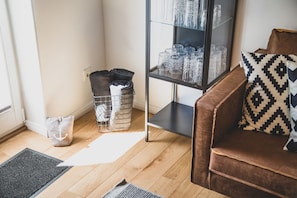 A living room with a brown sofa, a patterned pillow, a glass display cabinet with glassware, a wire basket with folded blankets, and a grey rug.