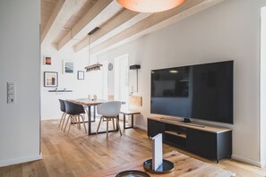 A modern living room with a large flat-screen TV, wooden flooring, and a dining area with chairs and a table.