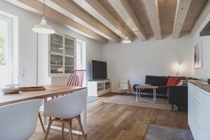 A modern living room with a wooden floor, a dining area with a wooden table and chairs, a black sofa, a TV, and a cabinet with glass doors.