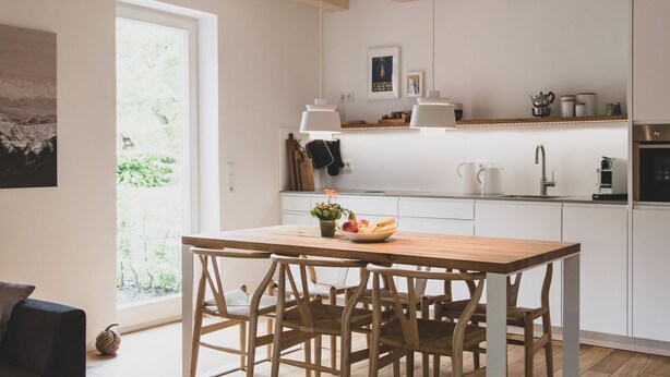 Una cocina moderna con armarios blancos, una mesa de comedor de madera y un amplio ventanal que proporciona luz natural.