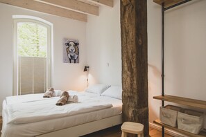 A bedroom with a large bed, a wooden beam, a window with blinds, a wall-mounted lamp, and a shelf with storage boxes.