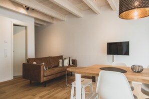 A modern living room with a wooden floor, a brown sofa with patterned cushions, a white dining table with chairs, and a flat-screen TV mounted on the wall. - Idyllische Fewo "jannes" - Gulfhof Boomgaarden (Krummhörn)