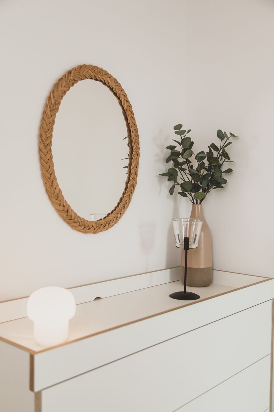 A white dresser with a woven mirror, a vase with green leaves, and a light bulb.