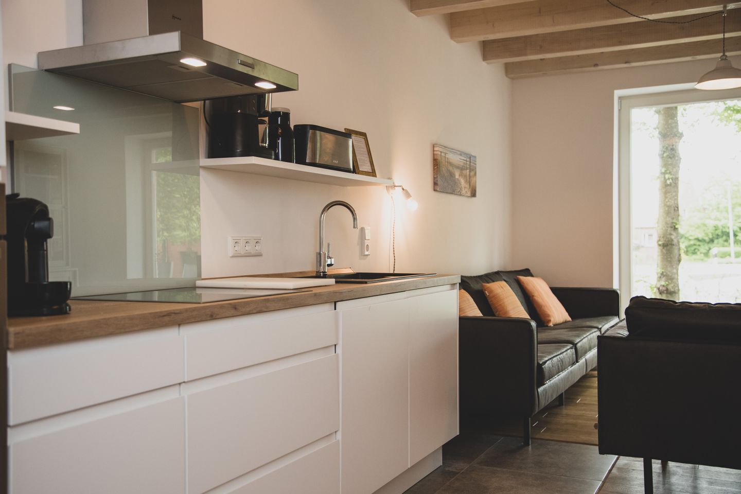 A modern kitchen with white cabinets, a wooden countertop, a black coffee machine, and a black sofa in the adjacent living area.