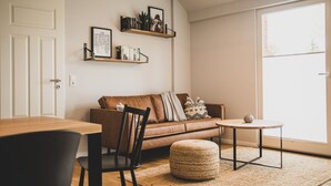 Ein gemütliches Wohnzimmer mit einem braunen Ledersofa, einem Holztisch und einem runden Ottomanen. Ein Bücherregal mit Büchern und Deko-Objekten ist vorhanden, sowie eine Tür links.