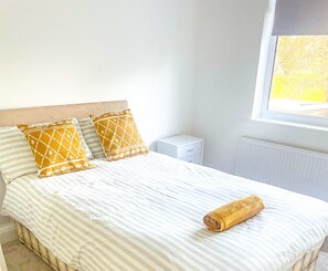 A neatly made bed with striped bedding and two patterned pillows, a white bedside table, and a window with a view of greenery. - 3-bed House in London Garden & Free Parking (Dartford)