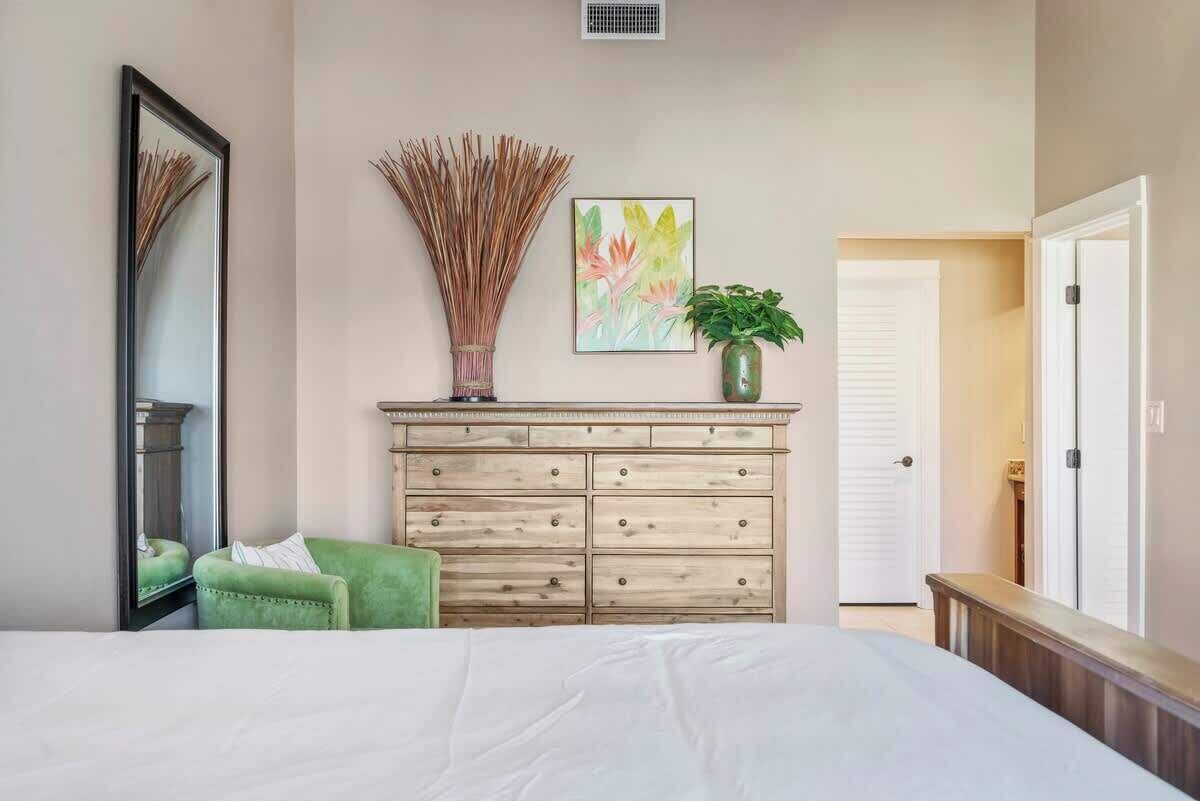 Een slaapkamer met een bed, een groene fauteuil, een houten dressoir, een spiegel, een vaas met gedroogde rietstengels en een schilderij.