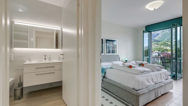 A modern bedroom with a large bed, a bathroom with a vanity, and a balcony with a view of the mountains. - Porto Letizia T4, Porlezza, Italy (Porlezza)