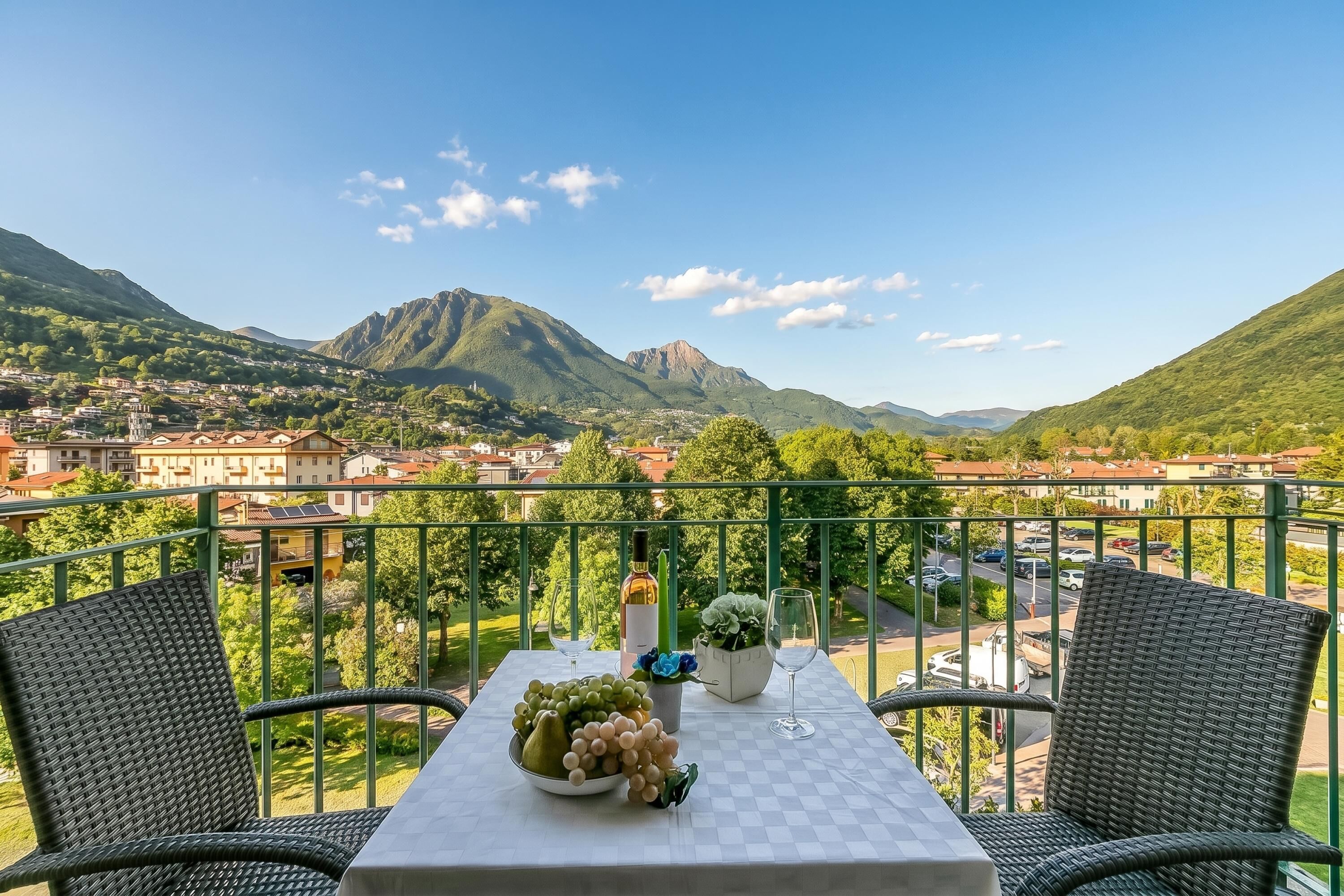 A balcony with wicker chairs, a table with a bowl of fruit, a wine bottle, and a glass. A mountain and town are visible in the background.