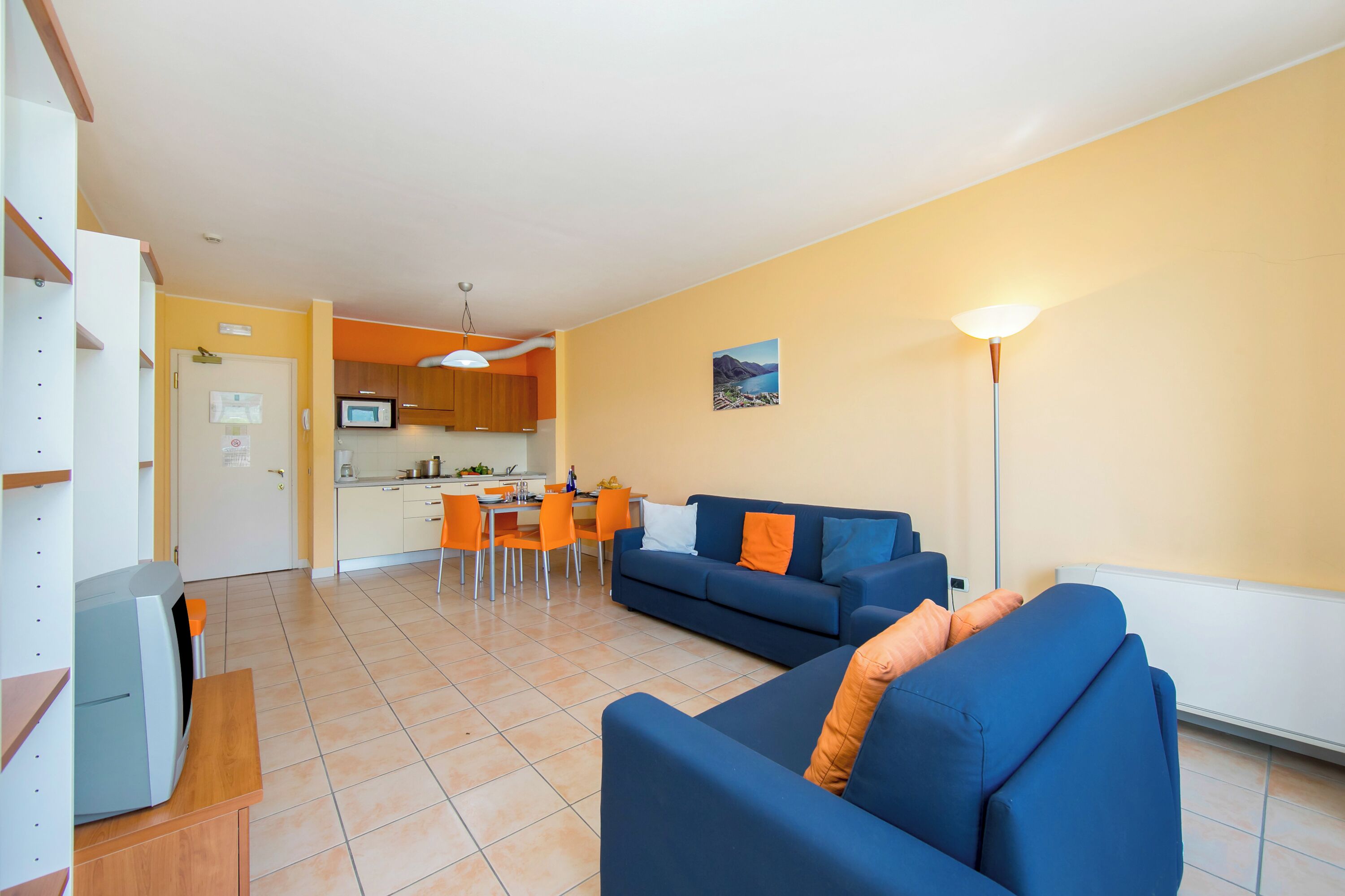 A living room with a blue sofa, orange cushions, a dining area with a table and chairs, and a kitchen in the background.