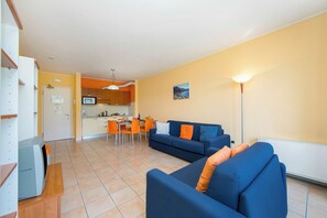 A living room with a blue sofa, orange cushions, a dining area with a table and chairs, and a kitchen in the background.