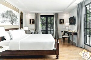 A hotel room with a large bed, a desk, a chair, and a view of trees through a window.