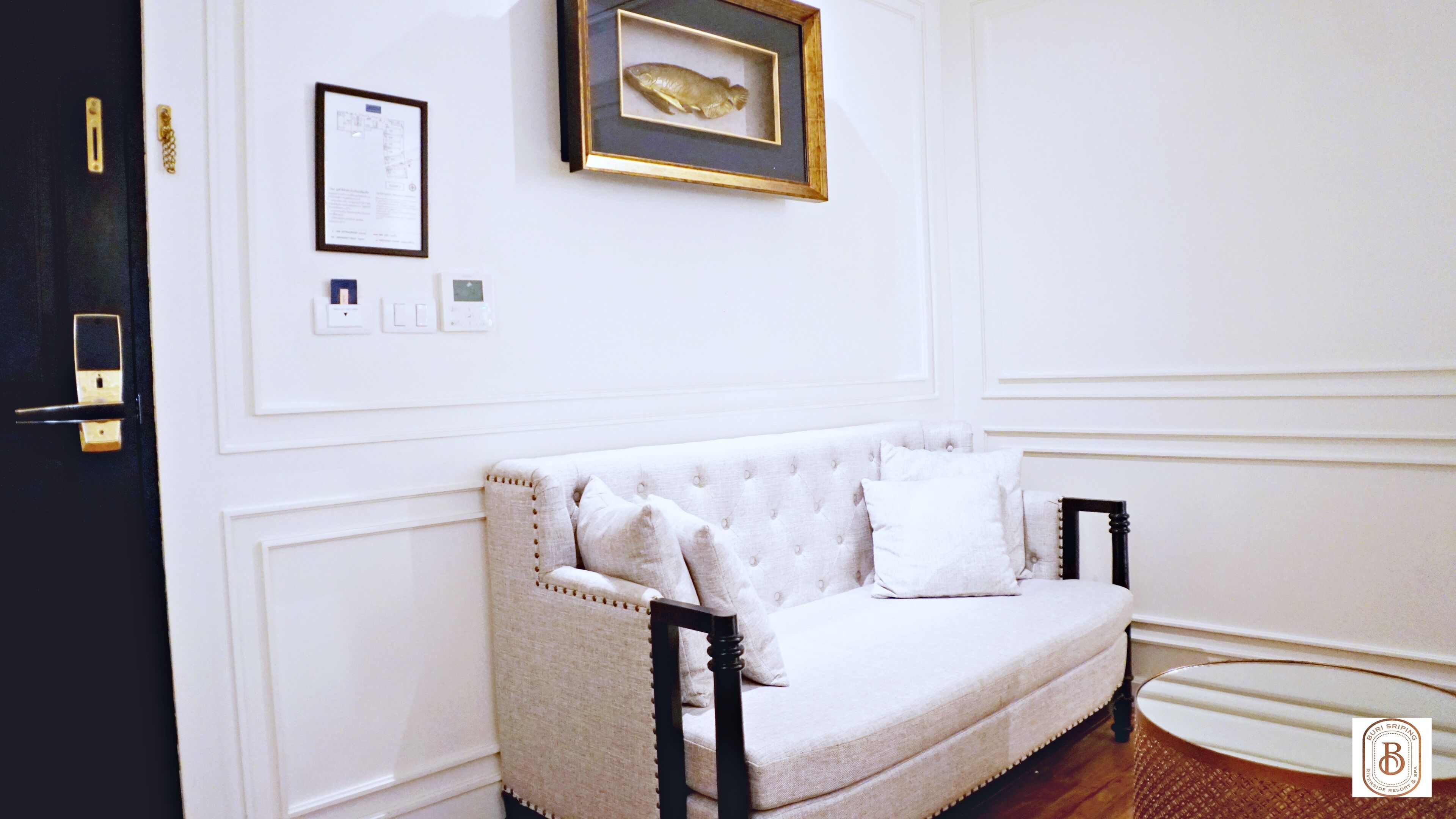 A white upholstered sofa with a tufted backrest and armrests, positioned against a white wall with a framed fish picture and a floor plan.