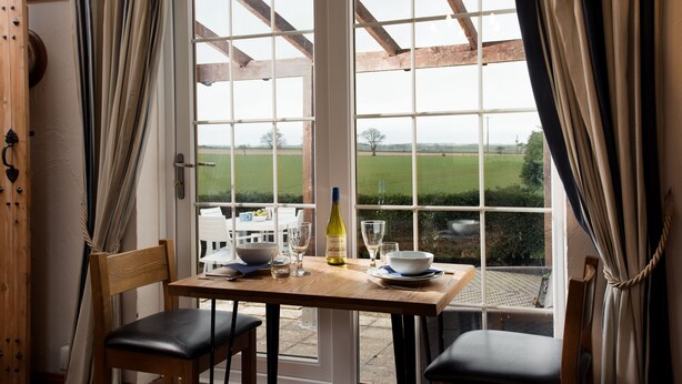 Ein Essbereich mit Tisch und Stühlen, einer Weinflasche und Blick auf ein grünes Feld durch ein großes Fenster.