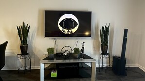Ein Wohnzimmer mit einem an der Wand befestigten Flachbildfernseher, einem Couchtisch mit elektronischen Geräten und zwei Blumentöpfen auf Beistelltischen.