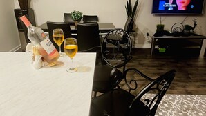 Ein Essbereich mit einem Tisch für zwei Personen, zwei Weingläsern und einer Weinflasche. Ein Fernseher an der Wand zeigt ein Programm.