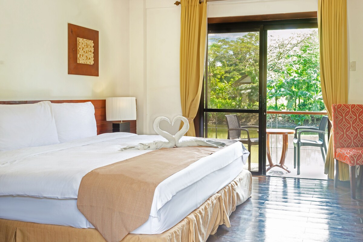 A hotel room with a large bed, a small table, a chair, and a balcony with a view of greenery.