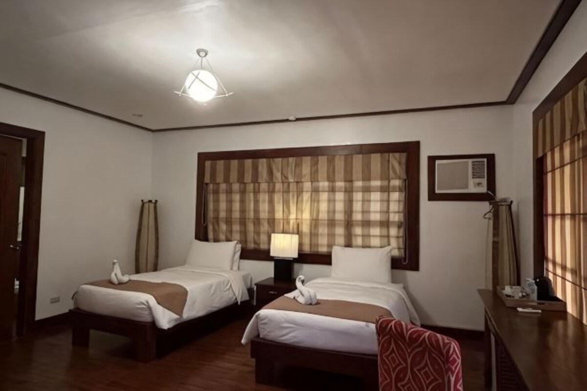 A hotel room with two beds, a nightstand, a lamp, a window with blinds, and a wall-mounted air conditioner.
