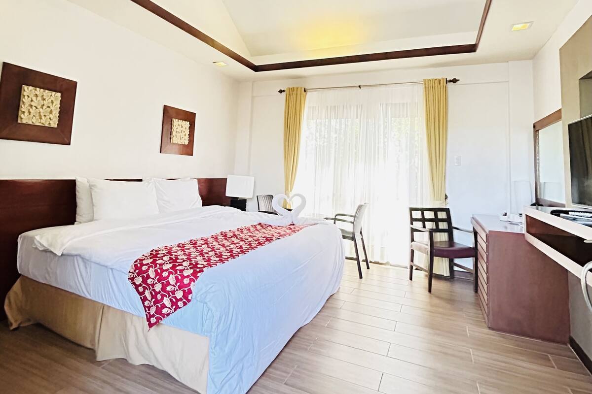 A hotel room with a large bed, a desk with a chair, a television, and a window with curtains.
