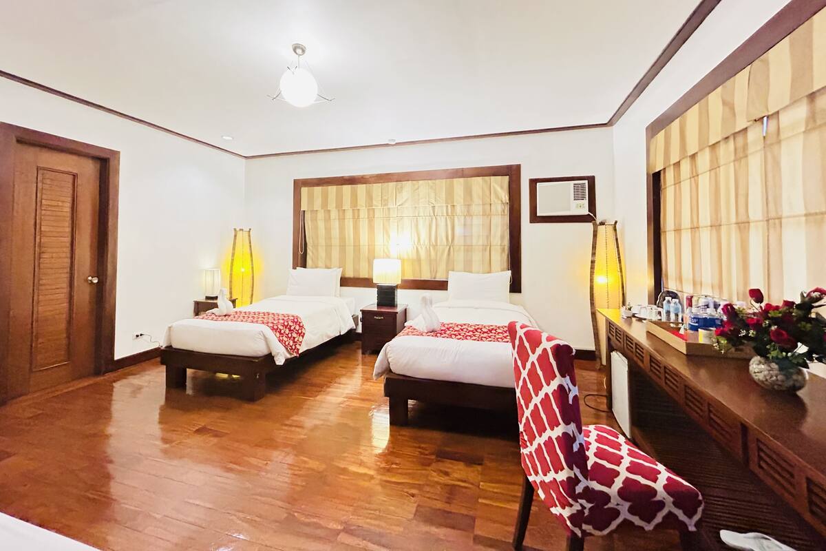 A hotel room with two beds, wooden flooring, a large window with curtains, a desk with a vase of flowers, and a chair.