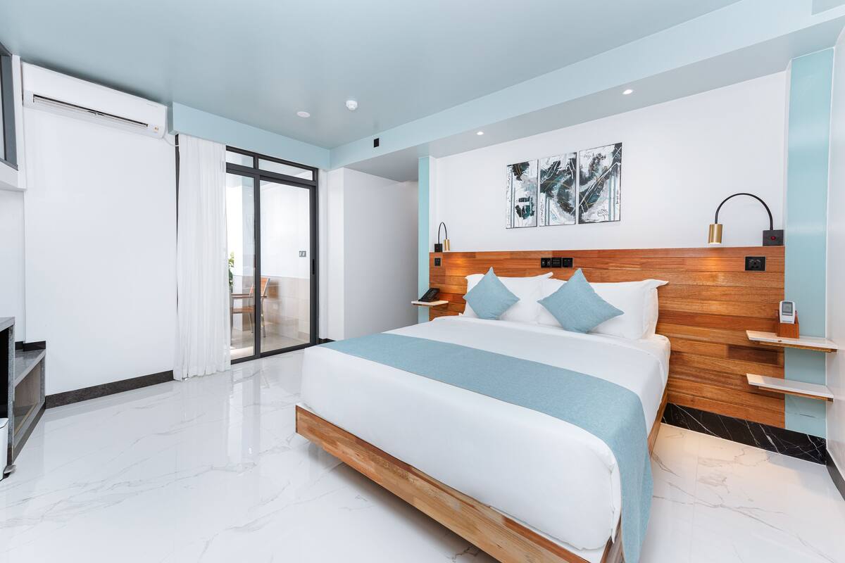 Une chambre d’hôtel moderne dotée d’un grand lit, d’une tête de lit en bois, d’une porte vitrée donnant sur un balcon et d’une œuvre d’art accrochée au mur.