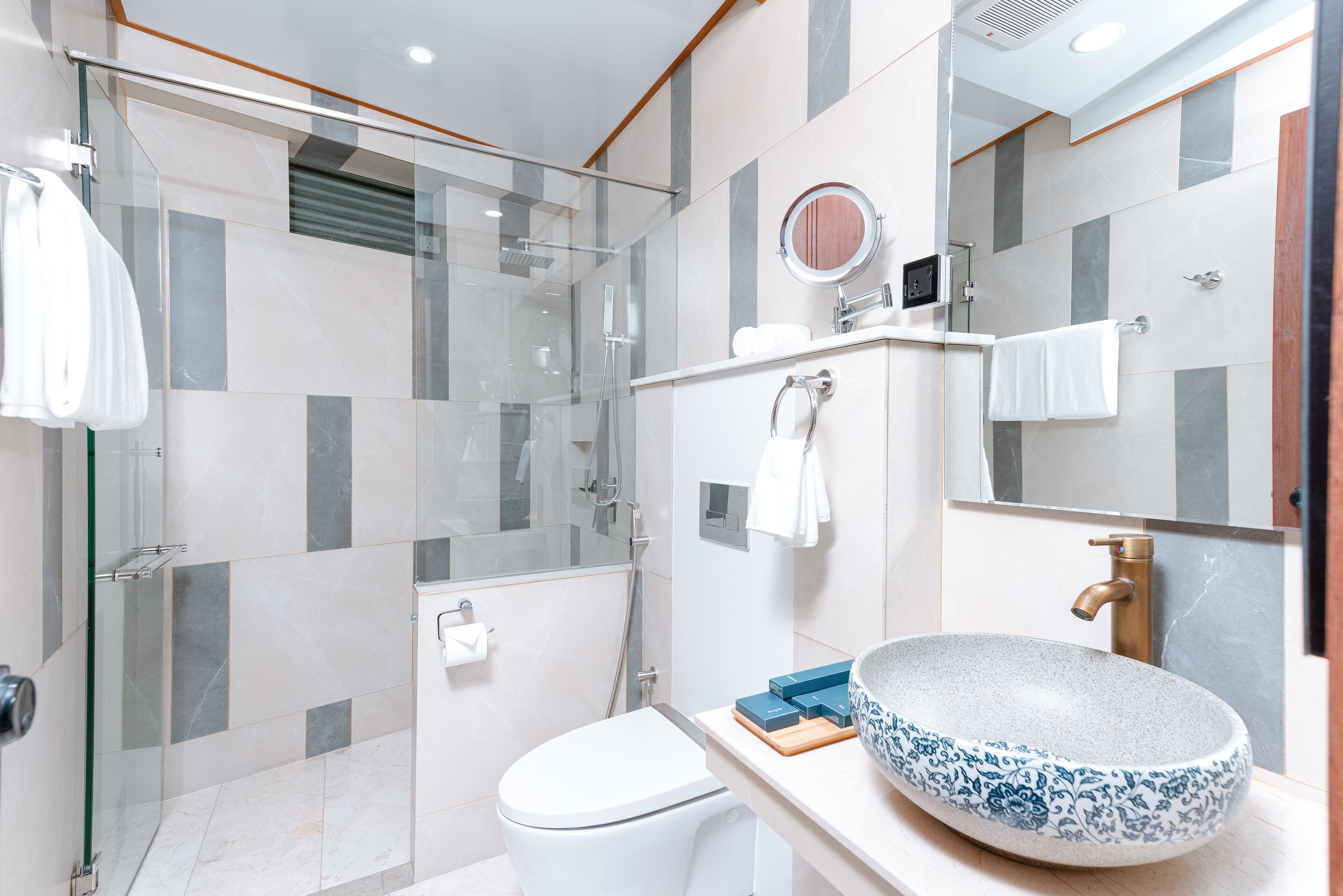 Un baño moderno con ducha de vidrio, inodoro blanco y un lavamanos grande con motivos florales azules.